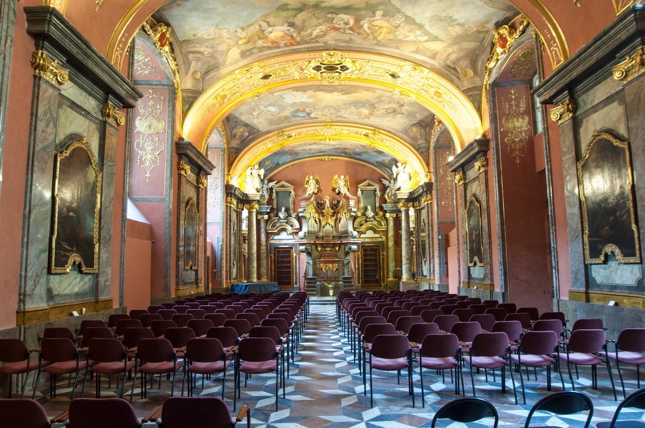 A long row of chairs in a chapel with colorful walls adorned with gold and a painting on the ceiling
