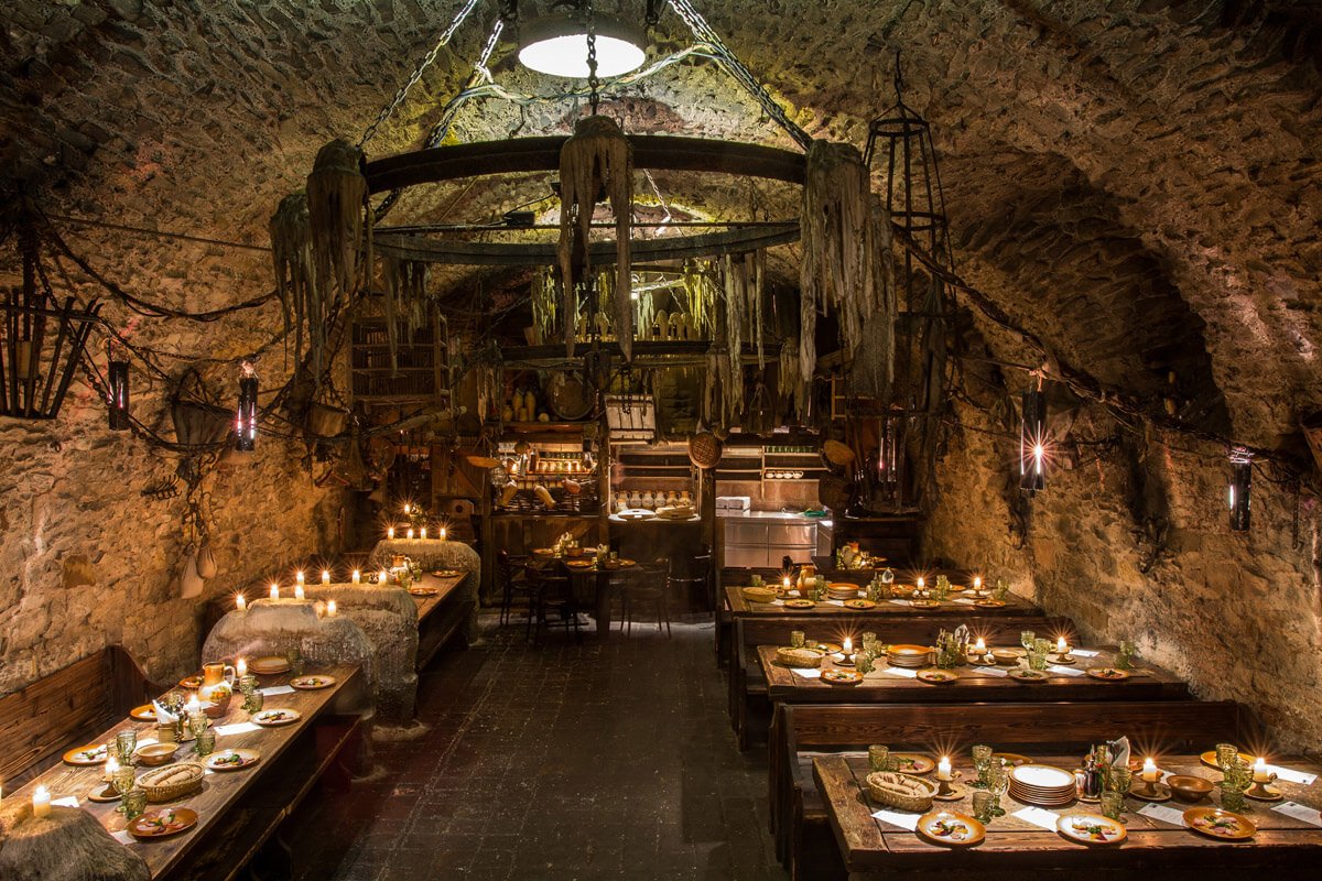 The interior of a medieval restaurant features a large chandelier in the center, the tables are lit by candlelight