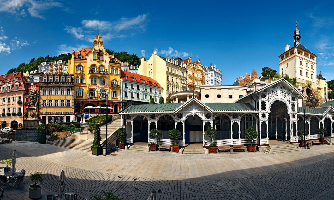 A square surrounded by tall, colorful buildings with a long white wooden gallery  carved with lace-like patterns