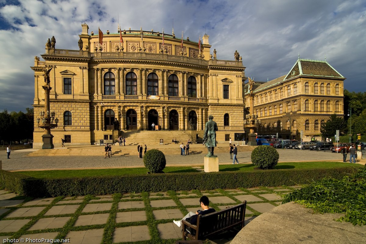 An imposing facade of a beige stone building,adorned with columns and statues with a statue in the square in front of it