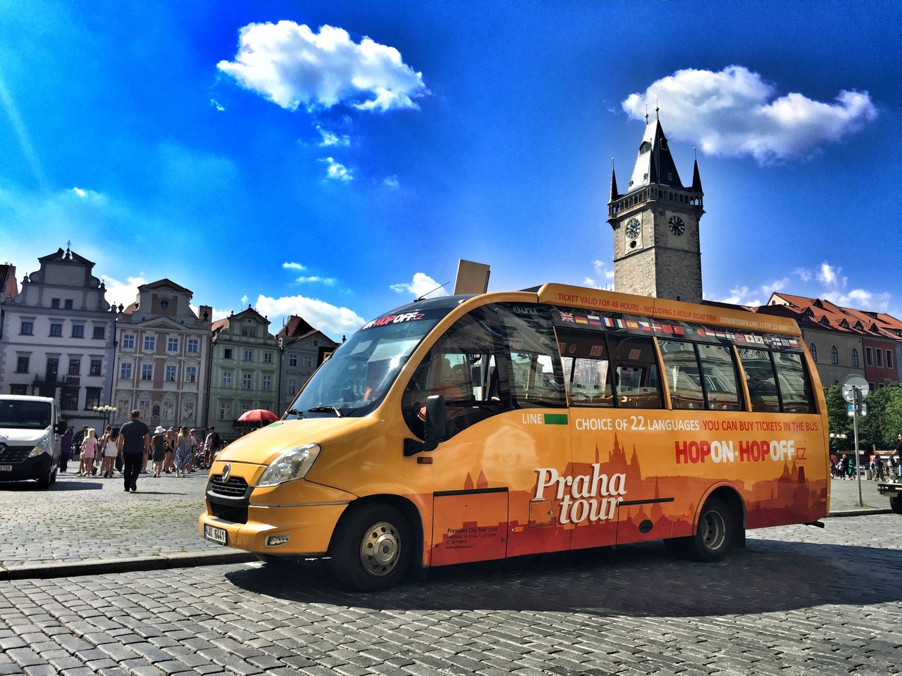An orange and yellow tourist bus is parked in a sunny square, in front of old buildings and a tall clock tower.