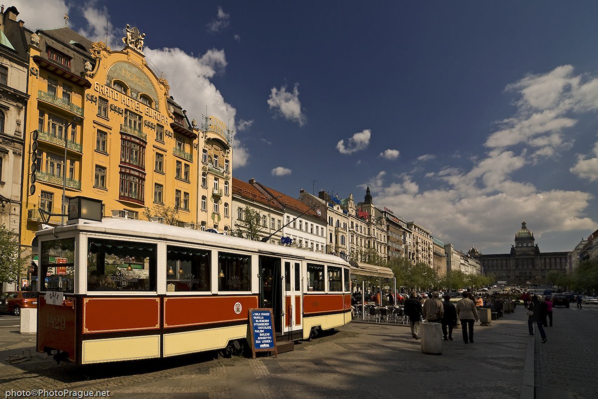 An old red and white tram is parked in a square lined with tall, colorful buildings, under a blue sky dotted with clouds
