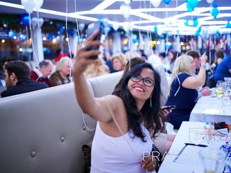 en:A woman taking a selfie in a crowd of people sitting on chairs; above them are blue and white balloons; fr:Une femme qui prend un selfie dans une foule assis sur des chaises, au-dessus il y a des ballons bleu et blanc