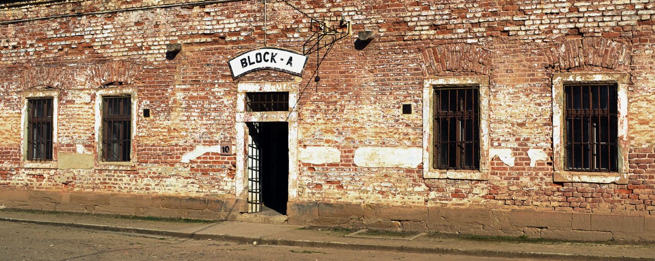 The facade of a building made of weathered red brick,with barred windows and an entrance bearing the inscription “BLOCK-A"