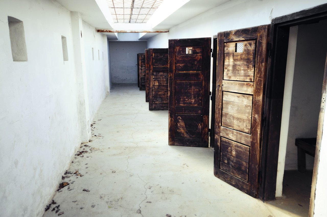 A white hallway with a rather dirty floor, lined with rooms whose wooden doors are badly damaged