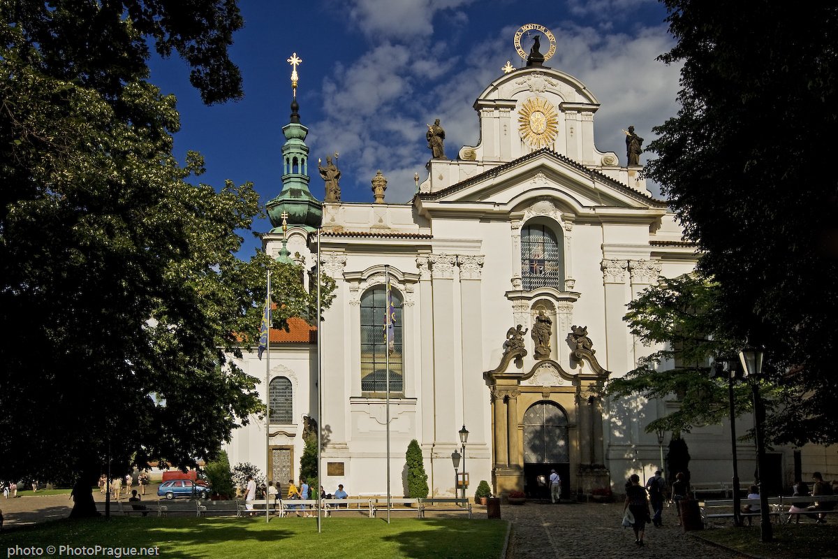 A white facade adorned with a statue and moldings, surrounded by trees and passersby under a blue, cloudy sky.