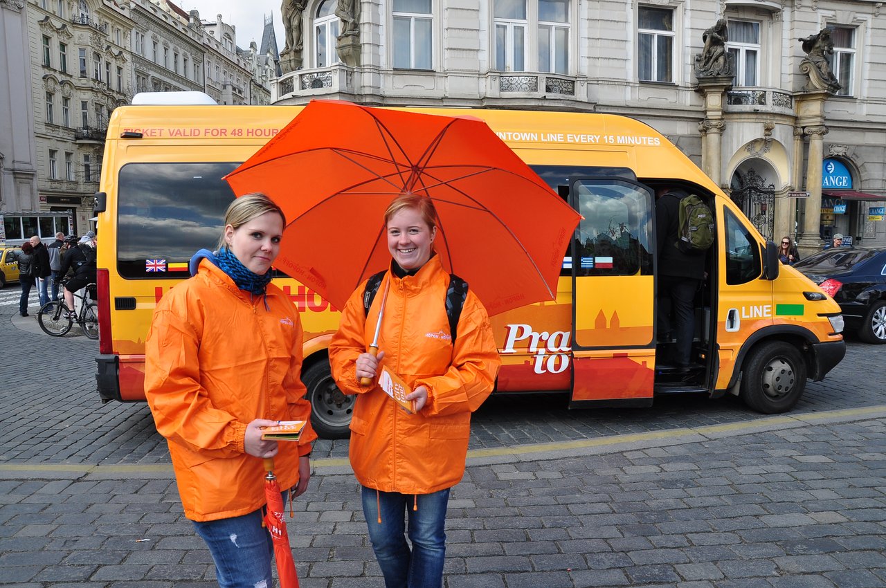 Two women dressed in orange and carrying orange umbrellas are handing out leaflets for the minibus