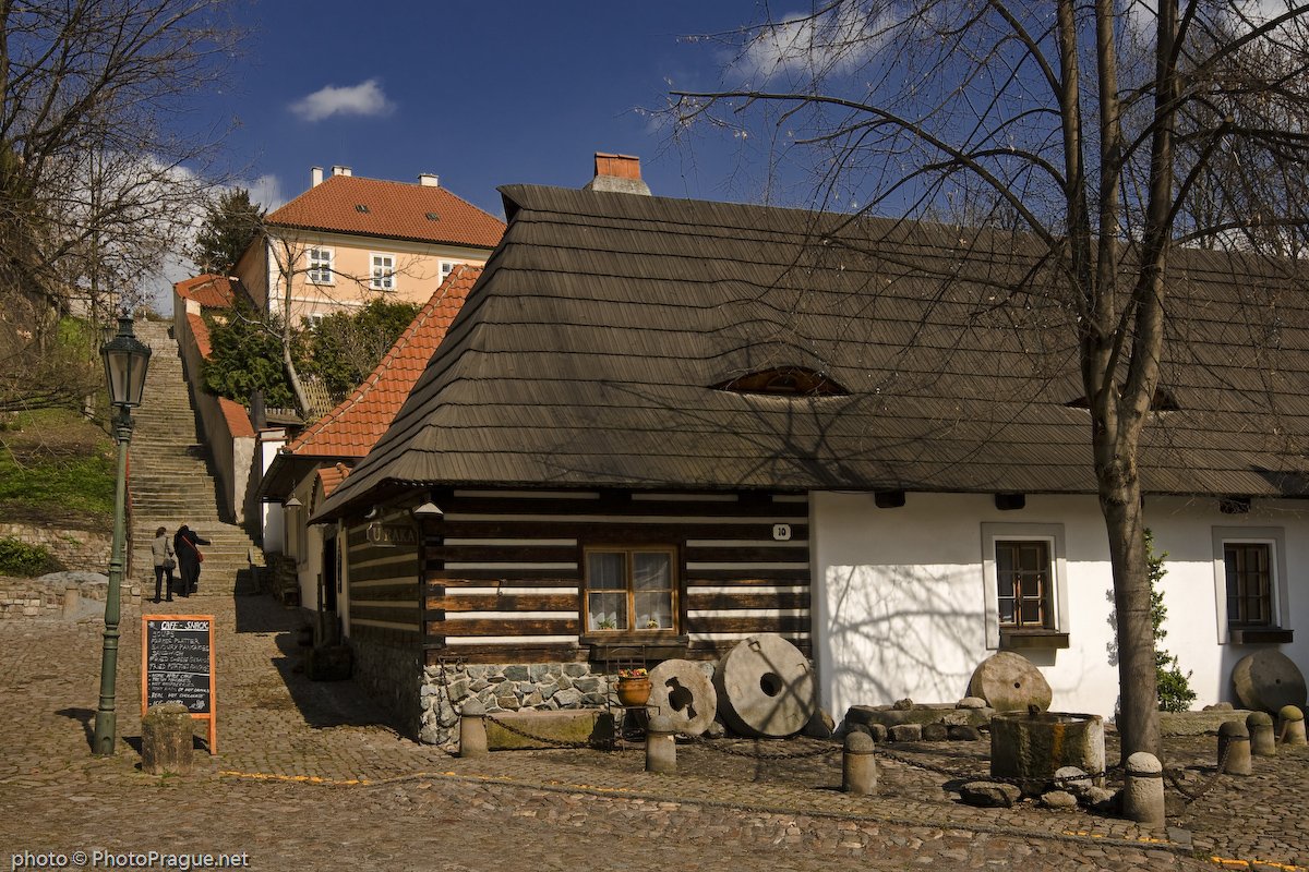 A cozy wooden house stands at the edge of a cobblestone alley under a blue sky dotted with clouds.